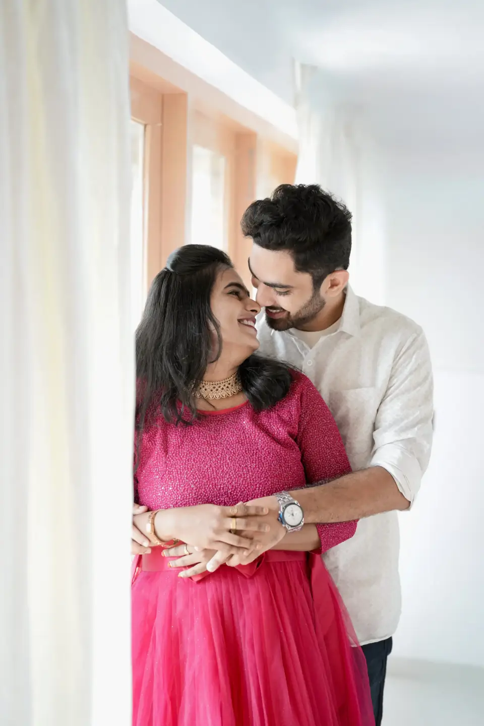 Husband kissing his wife on the cheek by a sunlit window, maternity shoot