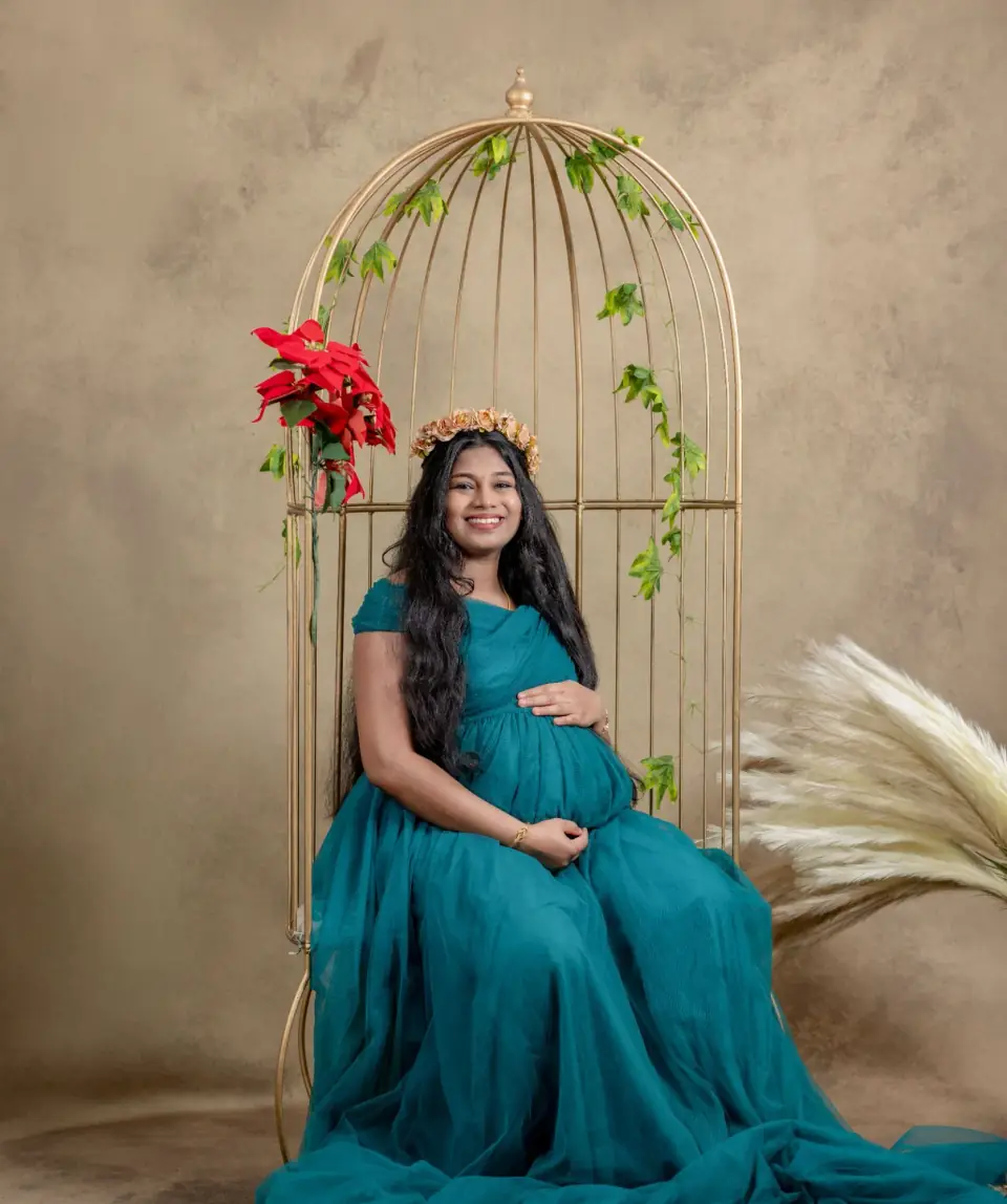 Mother-to-be in a teal gown seated in a golden birdcage set, studio maternity portrait